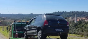 Guincho plataforma transportando um veículo Volkswagen preto em Telêmaco Borba, com fundo de vegetação e céu claro.