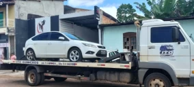 Guincho tipo plataforma transportando um carro branco, estacionado em Valença, próximo a um estabelecimento comercial.
