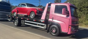 Guincho plataforma rosa transportando um carro antigo vermelho em Caxias do Sul, estacionado na beira da estrada.