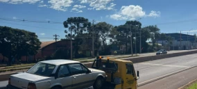 Guincho amarelo com veículo branco sendo transportado em Campo Mourão, em uma via com árvores ao fundo.