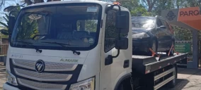 Guincho plataforma branco transportando um carro preto em Itapetinga, estacionado em um local com vegetação.