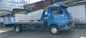 Guincho plataforma azul estacionado em Sapeaçu, com fundo de paredes brancas e céu claro.